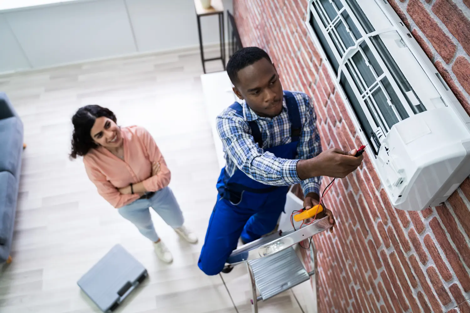 Young Woman Looking At Male Technician Repairing Air Conditioner Mounted On Brick Wall
